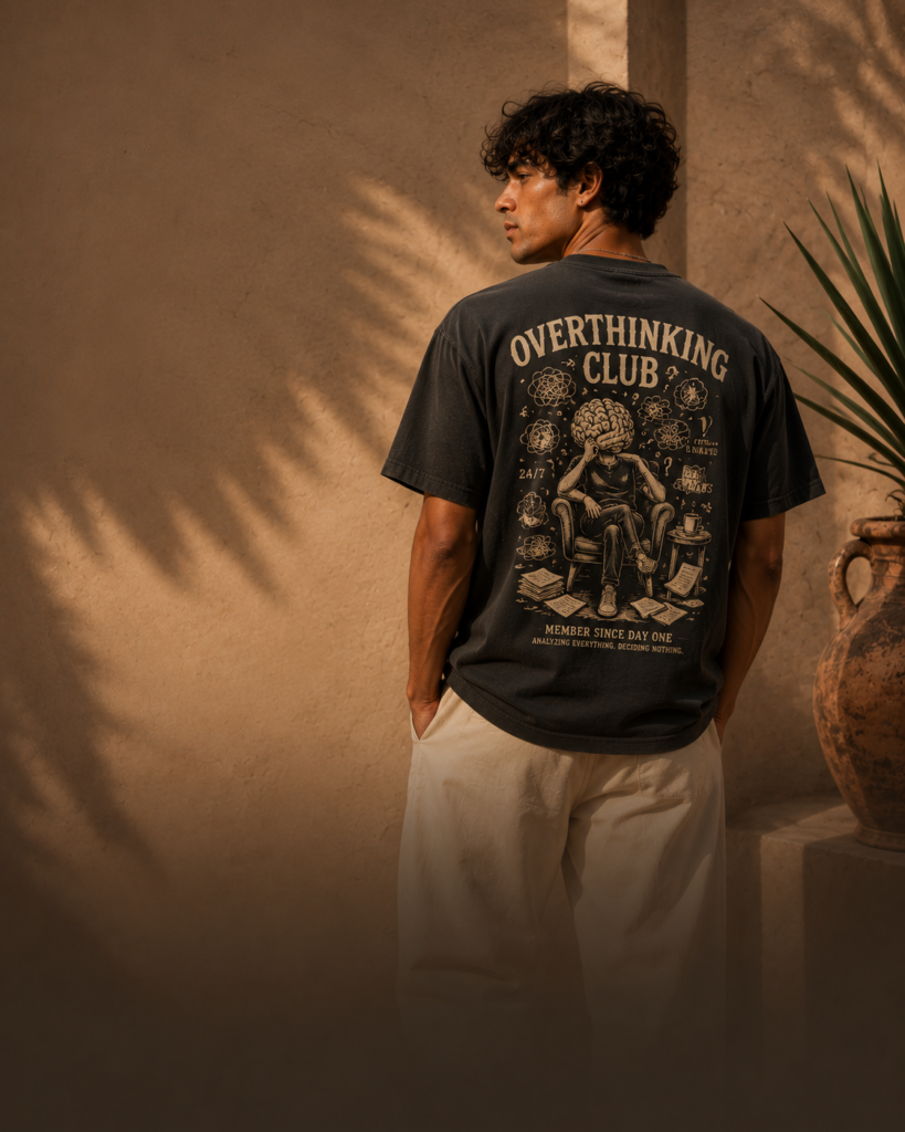 Adult male model wearing the Overthinking Club graphic T-shirt in a warm streetwear editorial setting.