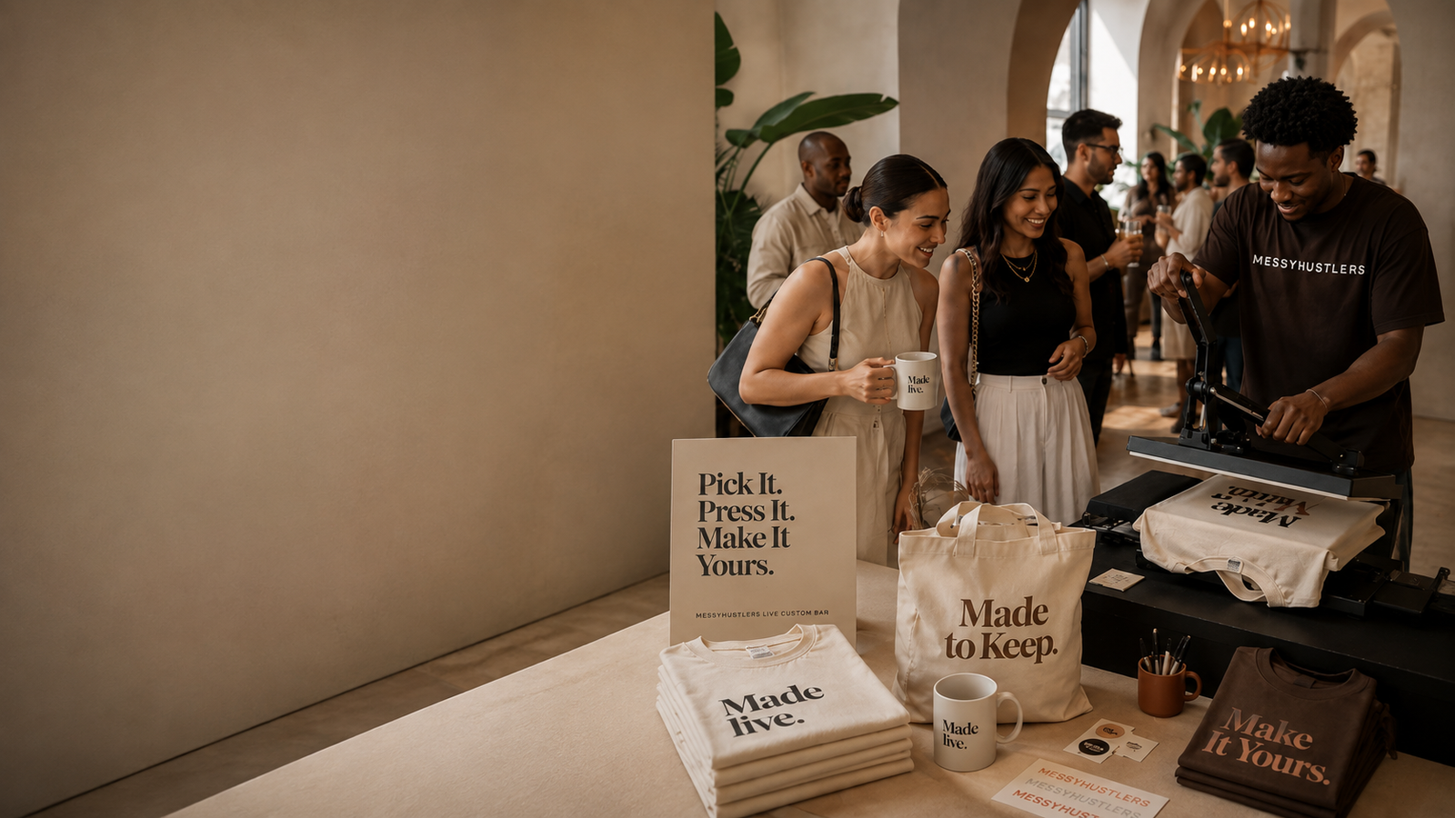 A live apparel customization booth with smiling guests, a team member at a heat press, folded shirts, a tote, and a mug in a warm event setting.